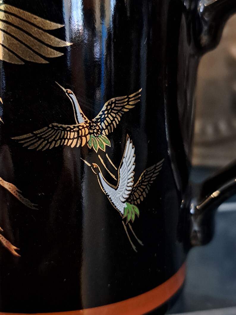 May include: Black ceramic mug with a gold and white crane design. The crane is flying with its wings spread. The mug has a brown rim.