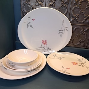 May include: A set of white ceramic dinnerware with a delicate floral design featuring pink roses and gray leaves. The set includes a large platter, a smaller dinner plate, a salad plate, and a bowl.
