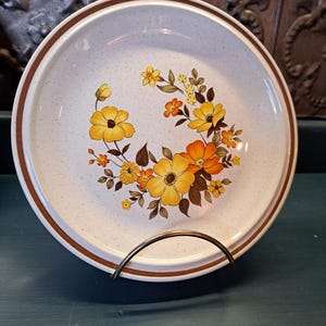 May include: A white ceramic dinner plate with a brown rim and a floral design featuring yellow, orange, and brown flowers. The plate has a speckled texture.