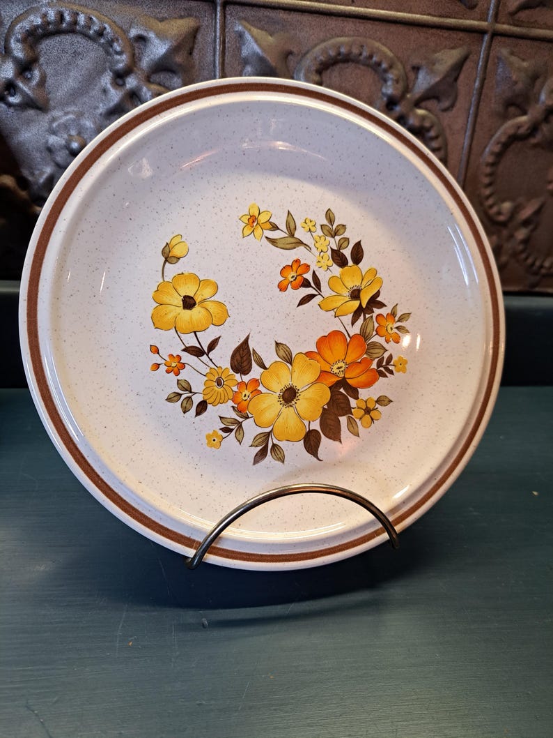 May include: A white ceramic dinner plate with a brown rim and a floral design featuring yellow, orange, and brown flowers. The plate has a speckled texture.