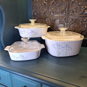 May include: A set of three white casserole dishes with glass lids. Each dish features a floral design with purple irises and green stems. The dishes are of varying sizes and are displayed on a teal surface.