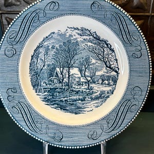 May include: A blue and white decorative plate with a landscape scene. The plate features a detailed illustration of a winter scene with trees, a river, and buildings. The rim has a patterned design with a dotted border.