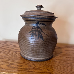 May include: A brown ceramic jar with a lid. The jar has a rounded body and a lid with a small knob. A dark blue leaf design is painted on the front. The jar is sitting on a wooden surface.
