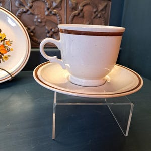 May include: A white teacup and saucer with a brown band around the rim. The cup has a speckled glaze and a rounded handle. The saucer has a matching brown band.
