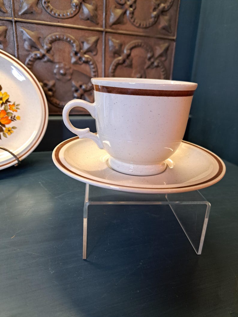 May include: A white teacup and saucer with a brown band around the rim. The cup has a speckled glaze and a rounded handle. The saucer has a matching brown band.