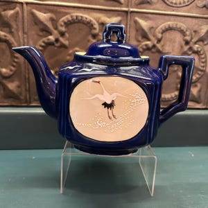 May include: A cobalt blue ceramic teapot with a square handle and spout. The teapot features a circular design with a crane illustration. The crane is white with black accents and a red head. The teapot is displayed on a clear acrylic stand.