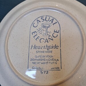 May include: A white ceramic plate with a blue and white logo that reads "Casual Elegance Hearthside Stoneware" and "Safe in your dishwasher, oven, & microwave oven." The plate also has the text "Crabli 572 Japan" printed on the bottom.