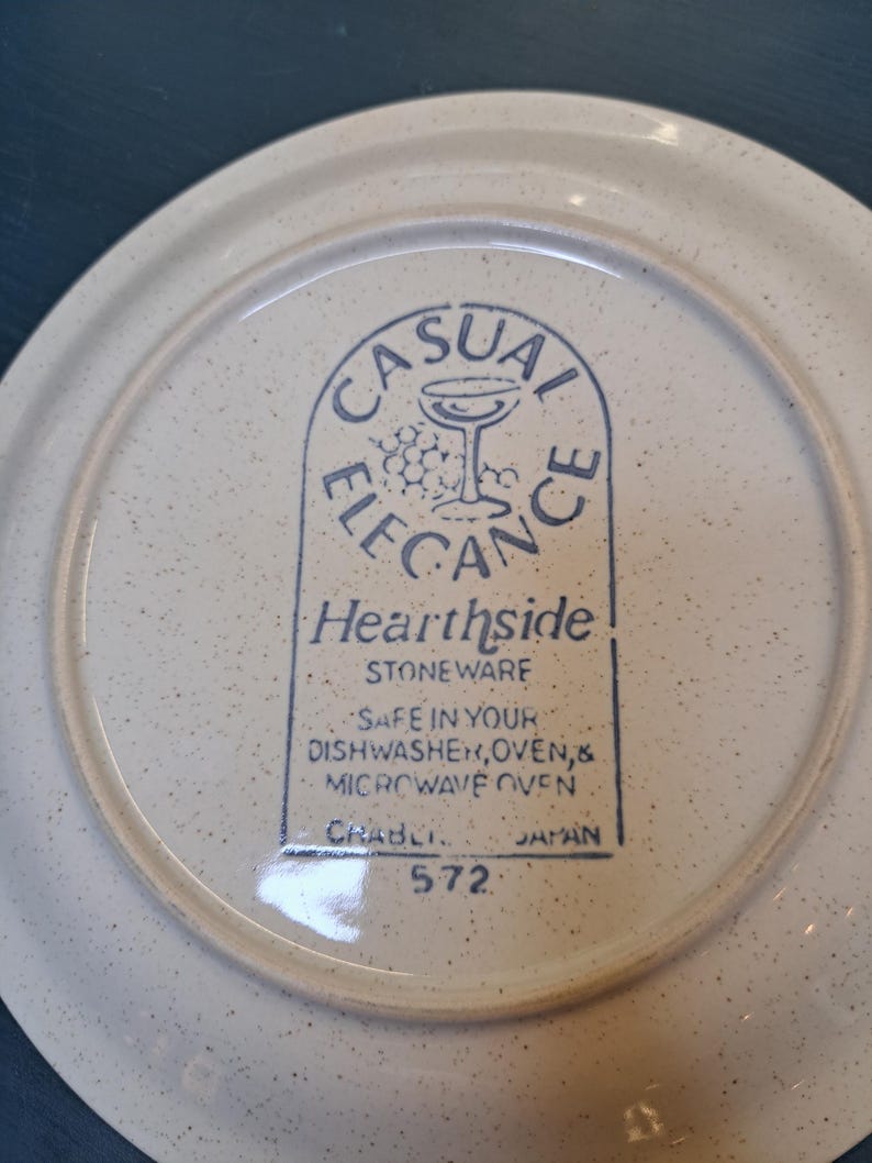 May include: A white ceramic plate with a blue and white logo that reads "Casual Elegance Hearthside Stoneware" and "Safe in your dishwasher, oven, & microwave oven." The plate also has the text "Crabli 572 Japan" printed on the bottom.