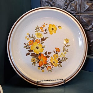 May include: A white ceramic dinner plate with a brown rim and a floral design featuring yellow, orange, and brown flowers.