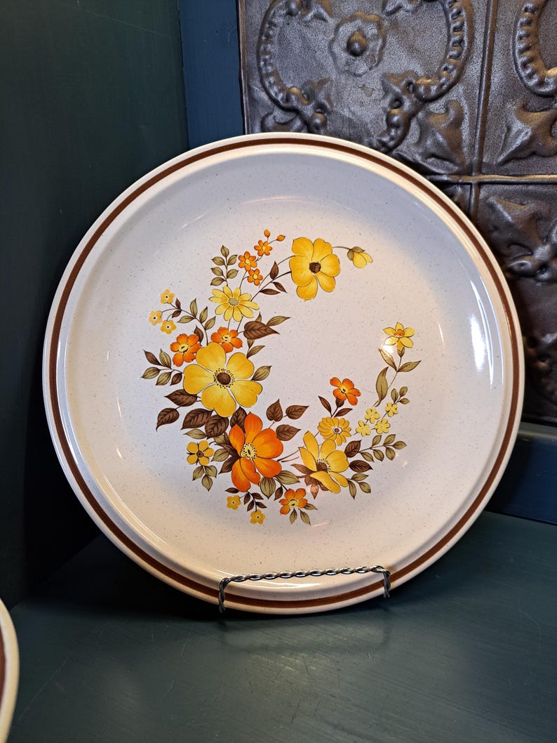 May include: A white ceramic dinner plate with a brown rim and a floral design featuring yellow, orange, and brown flowers.