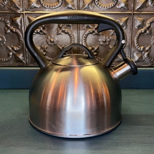 May include: A silver stainless steel kettle with a black handle and a black whistle. The kettle has a rounded body and a curved spout. The brand name "Cuisinart" is printed on the handle.
