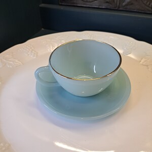 May include: A light blue teacup and saucer set with a gold rim. The cup and saucer are on a white plate with a floral design.