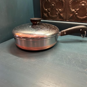 May include: A small, stainless steel saucepan with a copper band and a black handle. The pan has a lid with a black knob. The cookware is sitting on a blue surface.