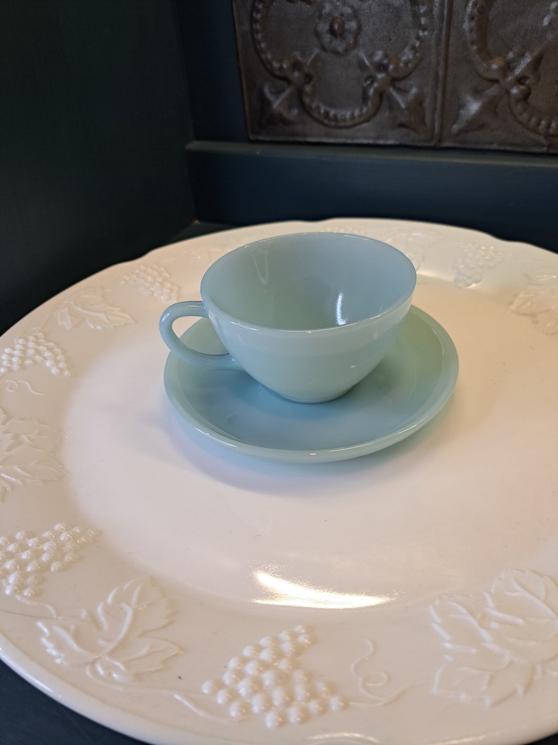 May include: A light blue teacup and saucer set on a white plate with a grapevine design.