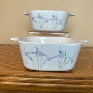 May include: Two white rectangular casserole dishes with handles, stacked. Each dish features a painted design of purple irises with green stems and leaves. The dishes are on a wooden surface.