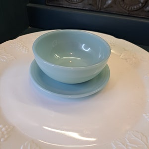 May include: A light blue glass bowl with a matching saucer. The bowl and saucer are sitting on a white plate with a grapevine design.