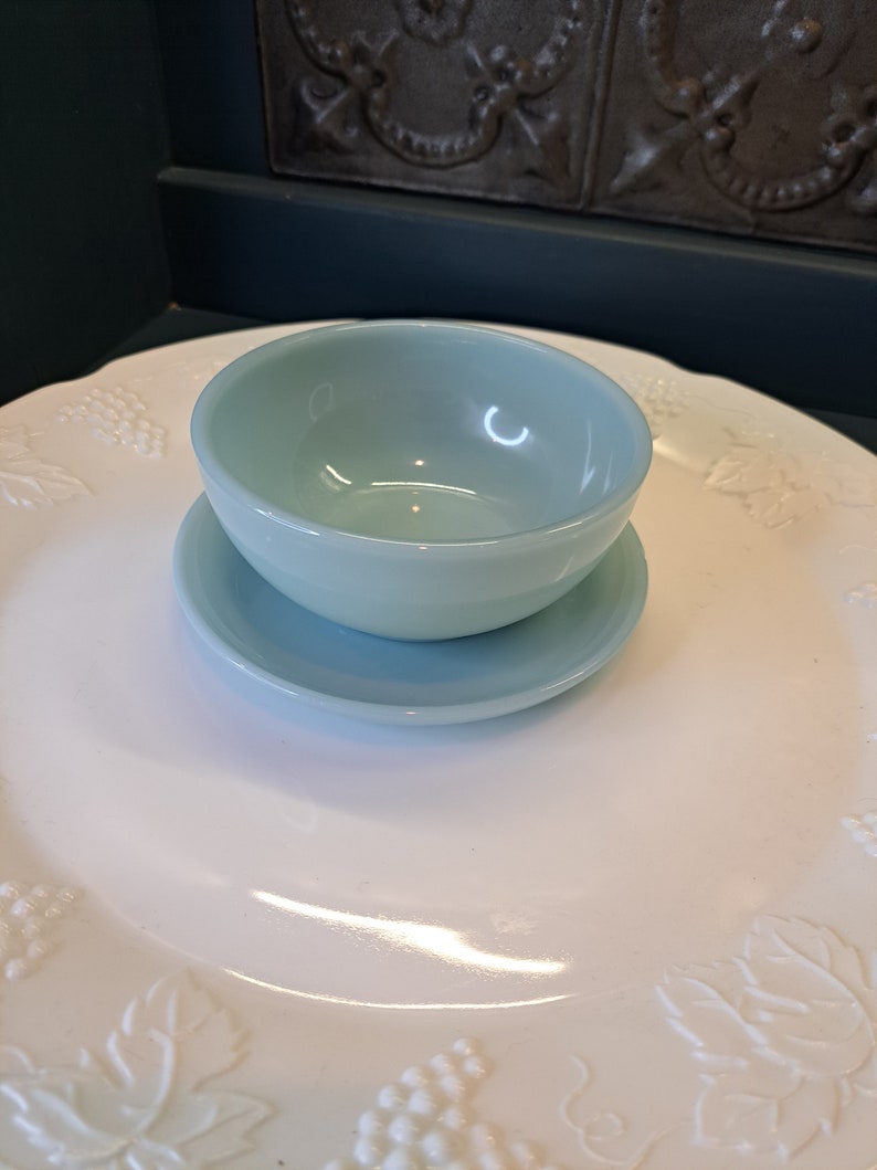 May include: A light blue glass bowl with a matching saucer. The bowl and saucer are sitting on a white plate with a grapevine design.