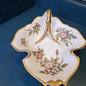 May include: A white porcelain serving dish with a gold handle and trim. The dish is leaf-shaped and decorated with pink and green floral patterns and a butterfly. The dish is approximately 8 inches long.