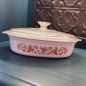 May include: An oval white casserole dish with a clear glass lid. The exterior of the dish features a horizontal band of colorful floral illustrations in shades of pink, yellow, and green, including small flowers and leaves encircling the base.