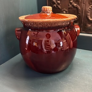 May include: A vintage, round, brown ceramic bean pot with a lid. The pot has two side handles and a glossy finish. The lid is a lighter shade of brown with a textured rim and a small knob.