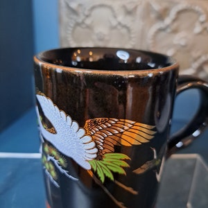 May include: Black ceramic mug with a gold rim and a design of white and gold cranes flying against a black background.