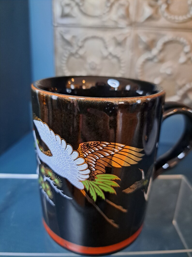 May include: Black ceramic mug with a gold rim and a design of white and gold cranes flying against a black background.