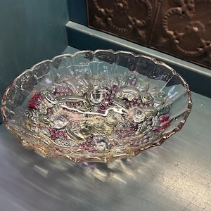 May include: A clear, oval-shaped glass bowl with a scalloped edge. The bowl features a decorative pattern of grapes and leaves in shades of purple and green. Three clear, round glass accents are evenly spaced within the bowl's design.