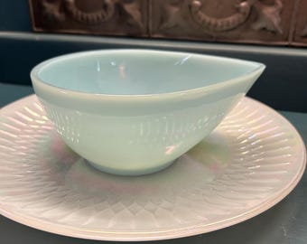 May include: A light blue, vintage-style gravy boat with a spout, resting on a matching iridescent saucer. The gravy boat has a ribbed texture. The saucer has a detailed, embossed pattern. The set is likely made of glass or ceramic.