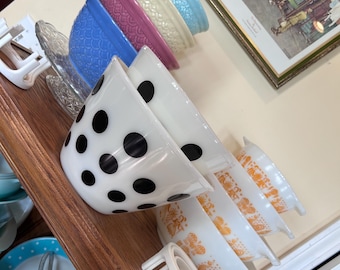 May include: A collection of vintage mixing bowls including a white bowl with black polka dots, a stack of orange patterned bowls, and a row of solid-colored textured bowls in blue, pink, cream, and light blue, arranged on a wooden shelf.