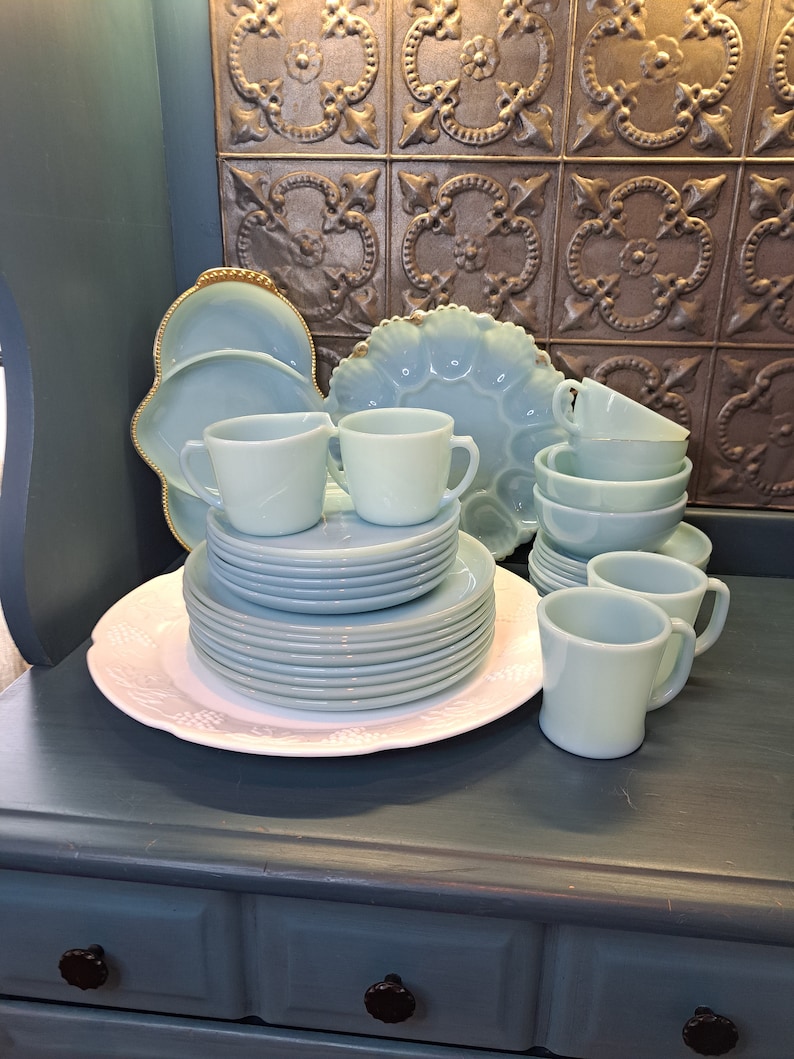 May include: A set of vintage blue milk glass dishes, including plates, bowls, mugs, and a serving platter. The dishes are arranged on a wooden surface with a decorative tile backsplash.
