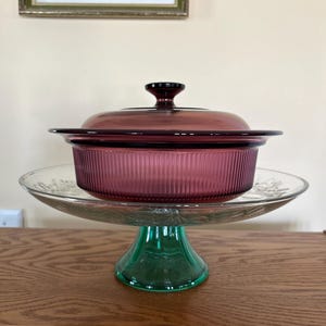 May include: A deep purple glass casserole dish with a matching lid featuring vertical ribbed sides, resting on a clear glass pedestal cake stand with a green base. The dish has a wide rim and a central knob on the lid for easy handling.