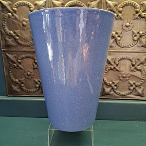 May include: A blue ceramic vase with a speckled texture. The vase has a wide opening and a slightly tapered shape. It is displayed on a clear acrylic stand. The background features a decorative metal panel.