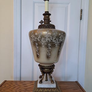 May include: A vintage table lamp with a gold-tone base and a white glass shade with a delicate lace pattern. The lamp has a marble base and a three-legged stand.
