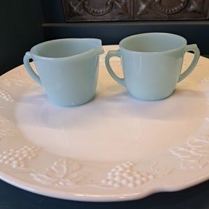 May include: A set of two light blue glass creamer and sugar bowls with handles. The bowls are on a white plate with a grapevine design.