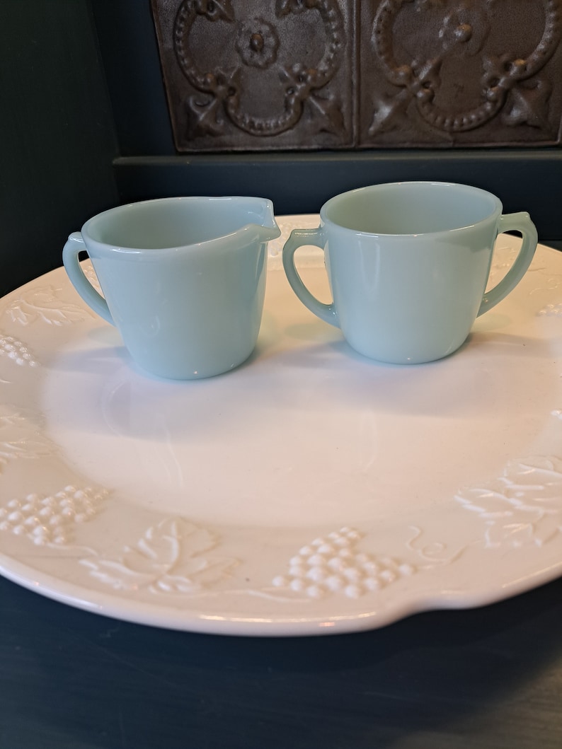 May include: A set of two light blue glass creamer and sugar bowls with handles. The bowls are on a white plate with a grapevine design.