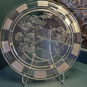May include: Clear glass plate with a floral rose design. The plate has a rectangular base and is supported by a clear plastic stand. The plate's rim features a repeating rectangular pattern.