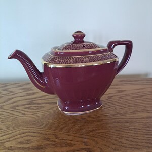 May include: A maroon teapot with a gold trim and a patterned lid. The teapot has a square body and a rounded spout.