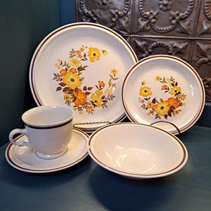 May include: A set of vintage floral dinnerware featuring a brown rim and a white background with a yellow, orange, and brown floral pattern.