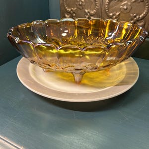 May include: An iridescent amber-colored glass bowl with a scalloped rim, resting on a white plate. The bowl has three small feet and a textured surface. The background features a decorative metal panel.