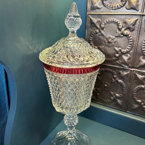 May include: A clear glass pedestal jar with a removable lid, featuring a diamond-cut pattern across the body and lid. A prominent horizontal band of deep red glass encircles the upper portion of the jar, which stands on a circular base.
