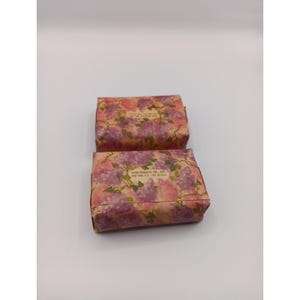 May include: Two rectangular bars of soap wrapped in floral paper. The paper has a pattern of pink and purple flowers on a beige background. The text "AVON PRODUCTS, INC., NYC" is visible on the packaging.