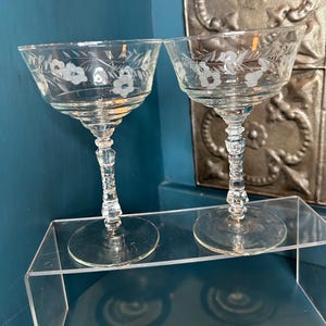May include: Two clear champagne coupe glasses with etched floral designs. The glasses have a wide bowl, a decorative stem, and a round base. The glasses are displayed on a clear acrylic shelf against a blue background.