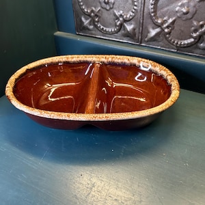 May include: Oval-shaped, two-sectioned brown ceramic dish with a glossy finish. The dish has a light brown, textured rim. The interior is divided by a central divider. This vintage serving dish is suitable for serving snacks or dips.