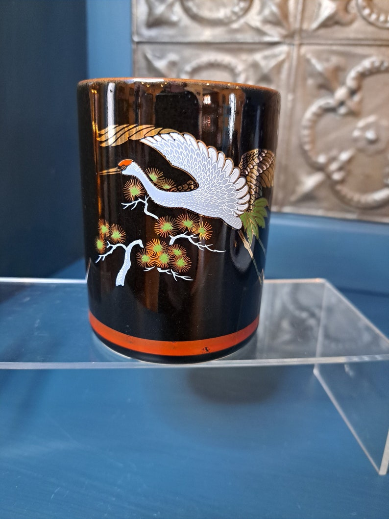 May include: Black ceramic cup with a gold and white crane design. The crane is flying over a branch with orange and green flowers.