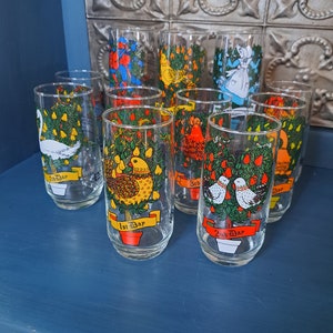 May include: Twelve glass tumblers with illustrations of the twelve days of Christmas. Each tumbler features a different day, with a colorful illustration of a bird, animal, or other symbol. The tumblers are arranged in a semi-circle on a blue surface.