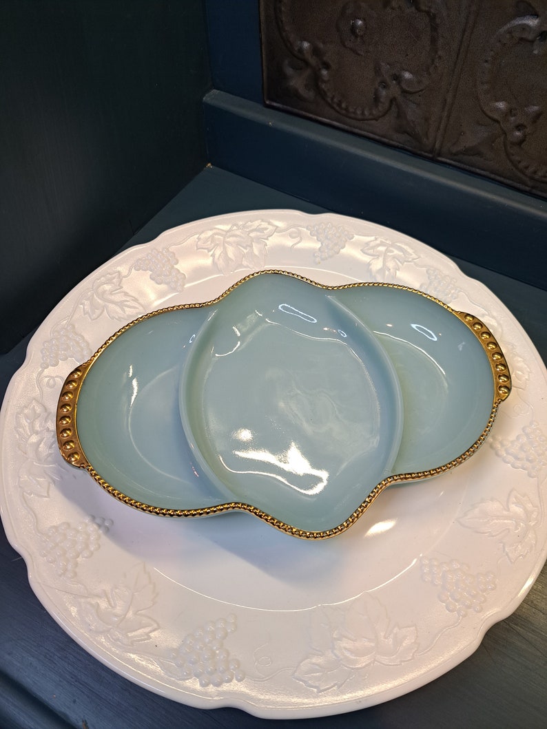 May include: A light blue glass serving dish with three sections and a gold rim. The dish is sitting on a white plate with a grapevine design.