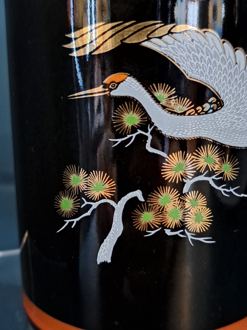 May include: Black ceramic vase with a white crane flying over a gold and green stylized pine tree design.