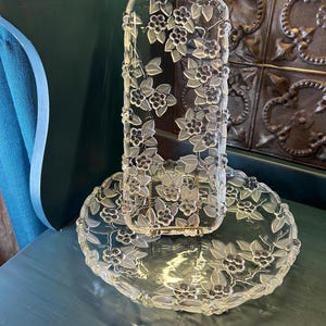 May include: Clear glass serving set featuring a floral design with frosted leaves and dark berry clusters. The set includes a rectangular holder and a round plate, both adorned with the same delicate pattern. The glass has a slight green tint.