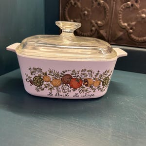 May include: A vintage, rectangular casserole dish with a clear glass lid. The dish is light pink with a decorative band of colorful vegetables and herbs. The text "Le Persil, La Sauge" is printed on the front.