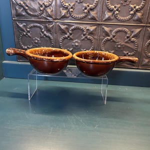May include: Two brown ceramic soup bowls with handles. The bowls have a glossy finish and a lighter brown rim. They are displayed on a clear acrylic stand against a textured, patterned background.